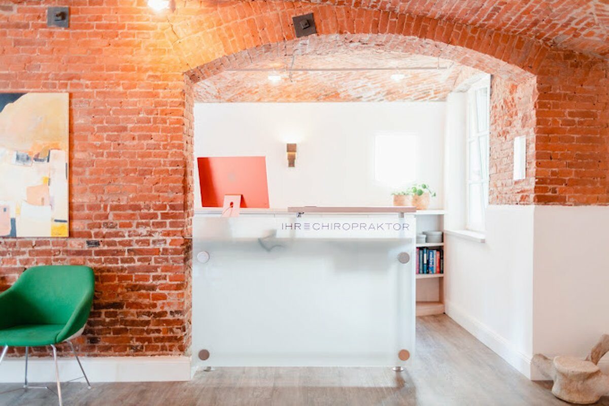 Heller Empfangsbereich einer Chiropraktik-Praxis mit Backsteinbogen, Glasrezeption, grünem Stuhl und Fensterlicht.