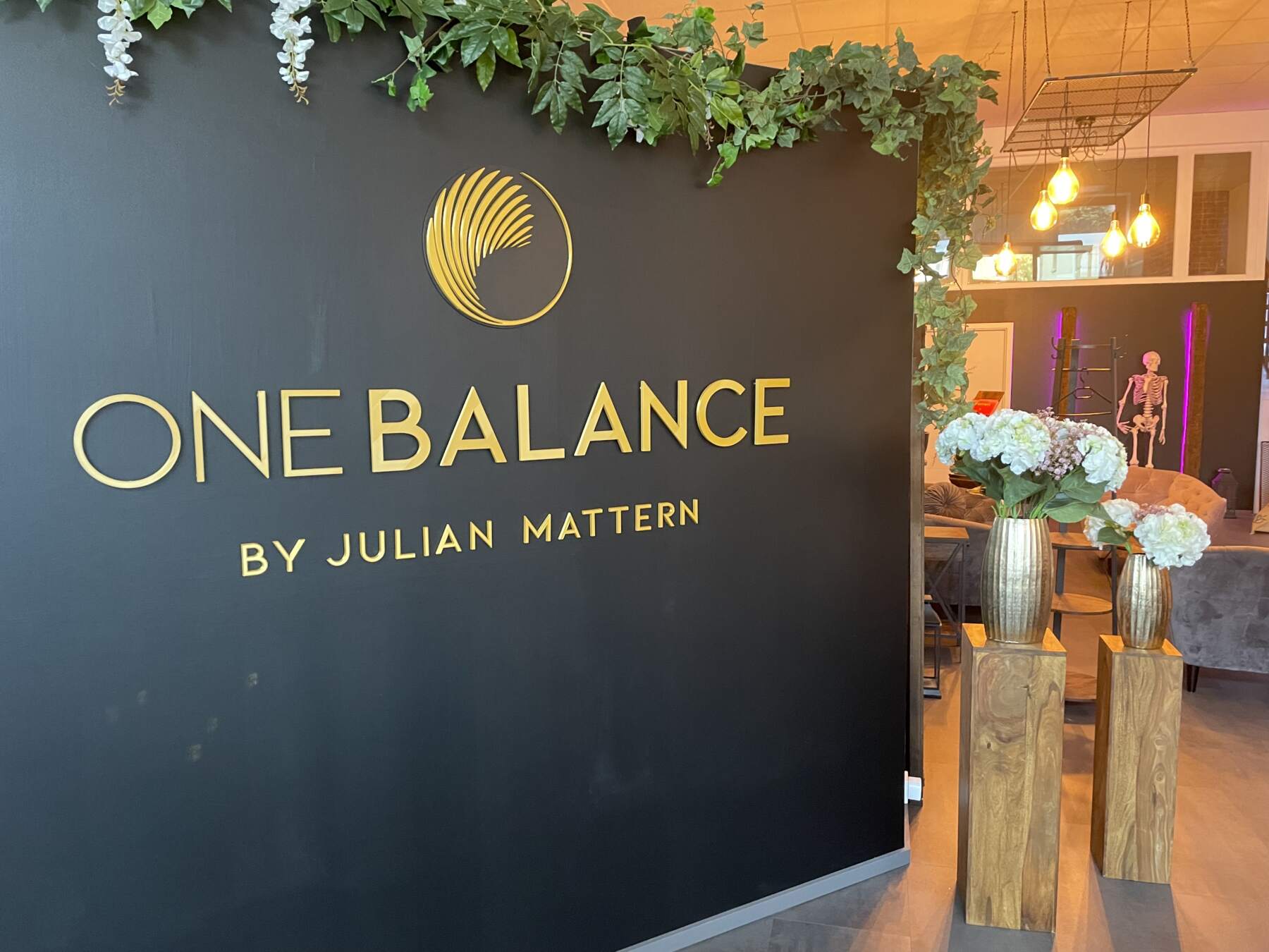 Schwarze Wand mit ONE BALANCE Schriftzug, Pflanzen und Vasen im modernen Empfangsbereich mit warmem Licht.