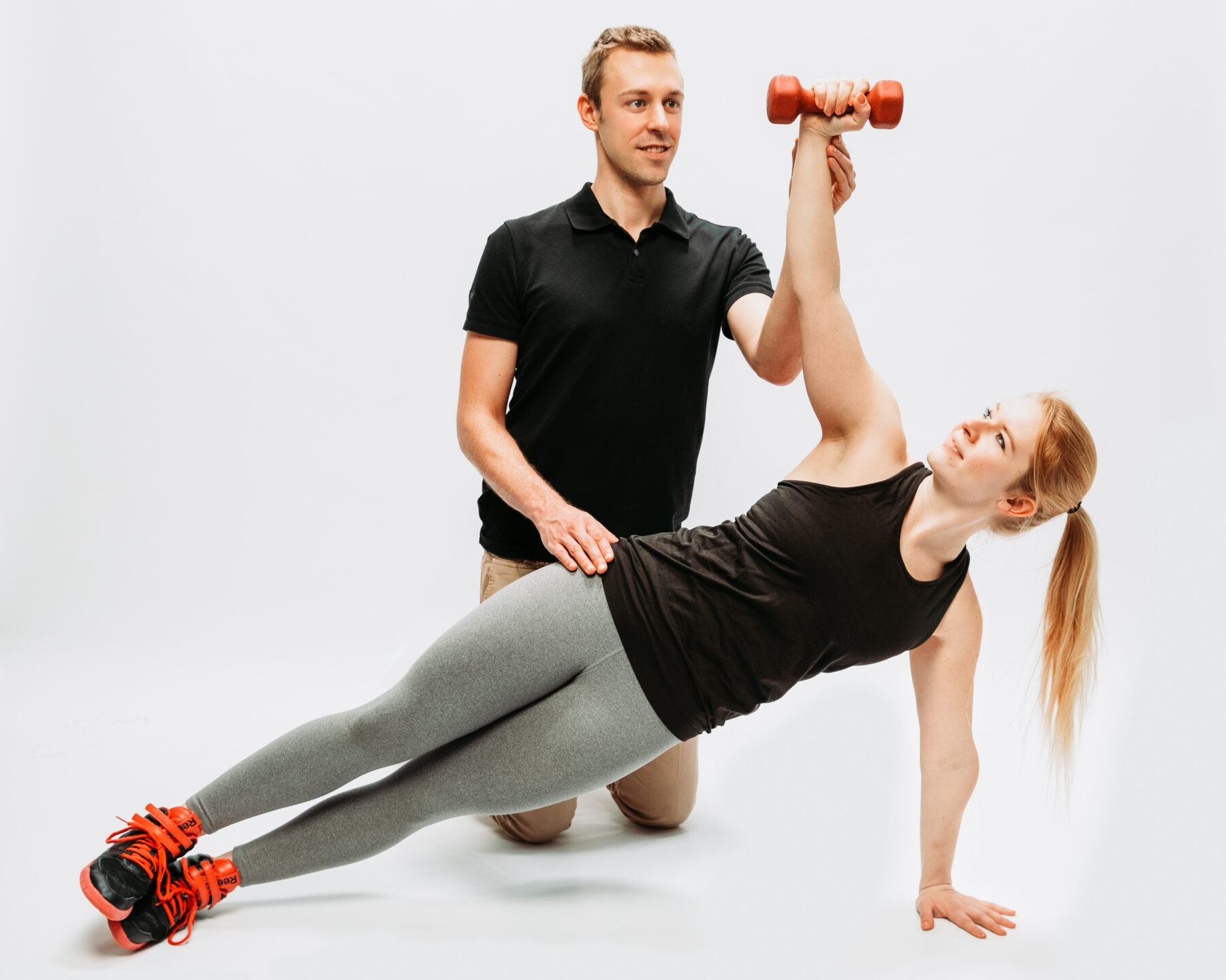 Personal Trainer betreut Frau beim Side-Plank und Schulterdrücken mit roter Kurzhantel im Fitnessstudio.