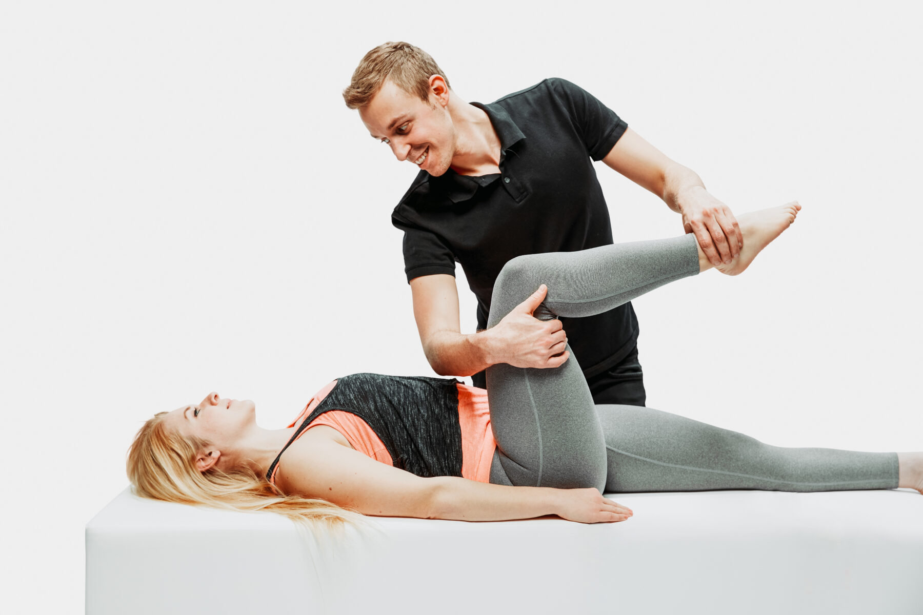 Physiotherapeut führt Dehnübung am Bein einer Patientin auf einer Behandlungsliege durch, Studioaufnahme vor weißem Hintergrund.