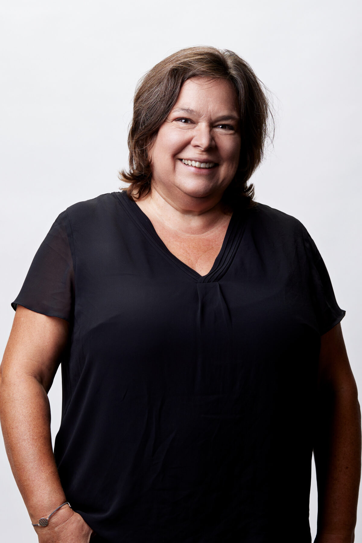 Lächelnde Frau mit schulterlangem braunem Haar in schwarzer Bluse vor hellem Studiohintergrund, Porträtaufnahme.