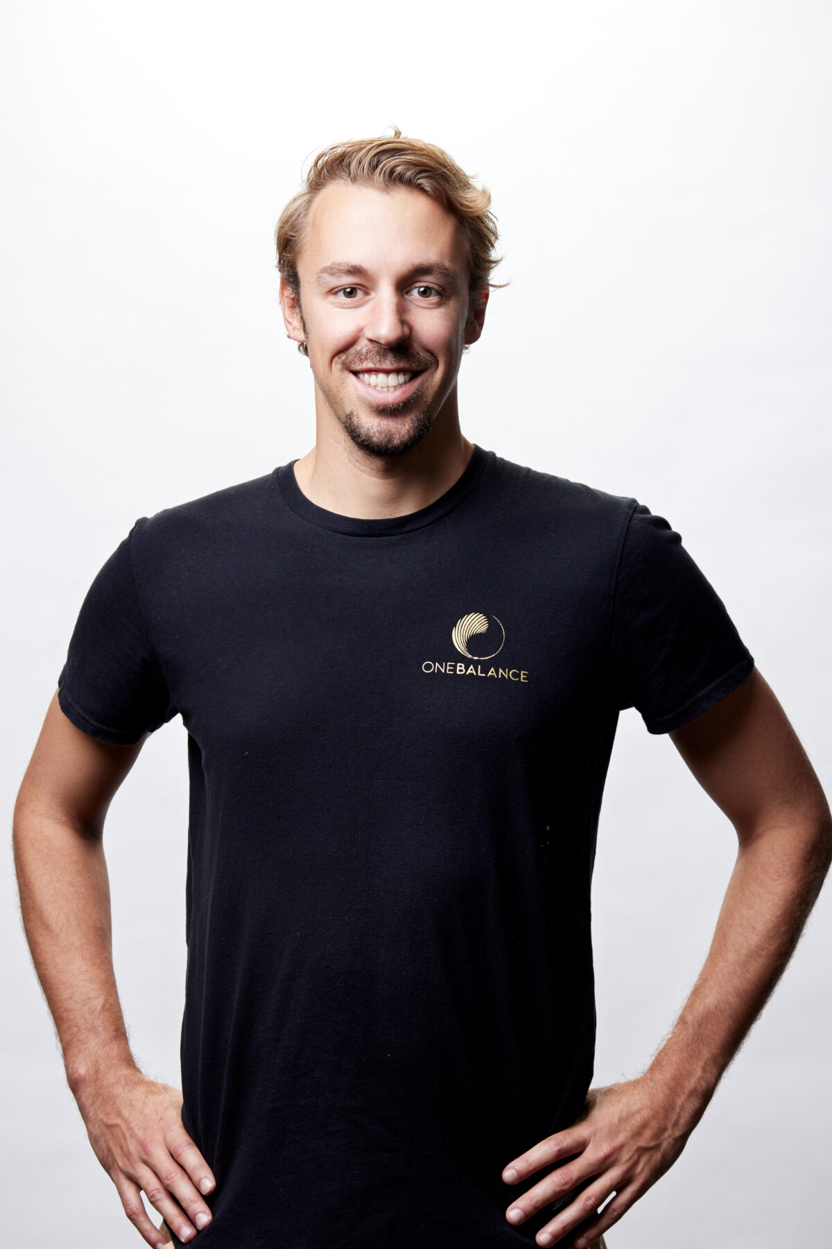Studio-Porträt eines lächelnden Trainers mit schwarzem T-Shirt und OneBalance-Logo vor hellem Hintergrund.