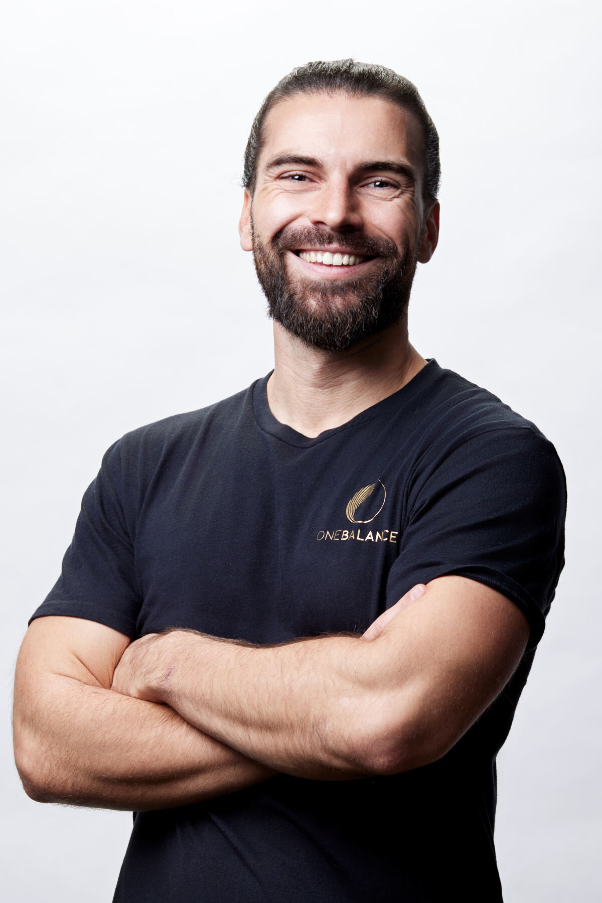 Lächelnder Mann mit Bart im schwarzen T-Shirt, Arme verschränkt, vor hellem Hintergrund.