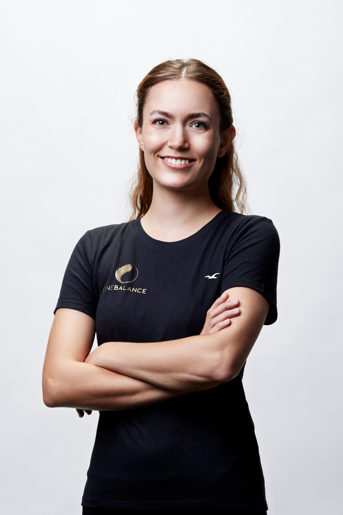 Lächelnde Trainerin mit verschränkten Armen vor hellem Hintergrund, trägt schwarzes Shirt mit Logo.