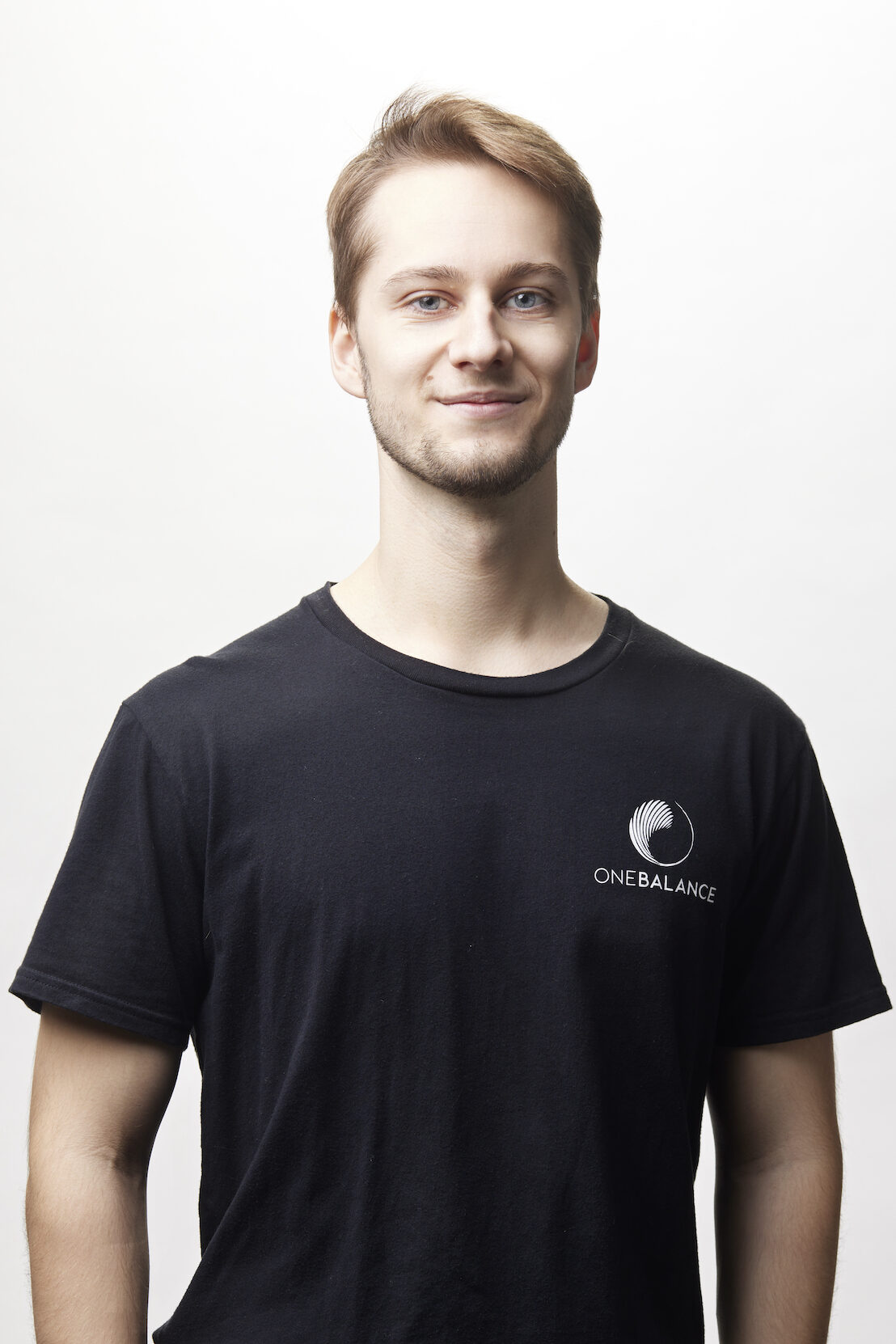 Studio-Porträt eines jungen Mannes mit kurzem Haar und leichtem Lächeln, schwarzes T-Shirt mit Logo vor weißem Hintergrund.
