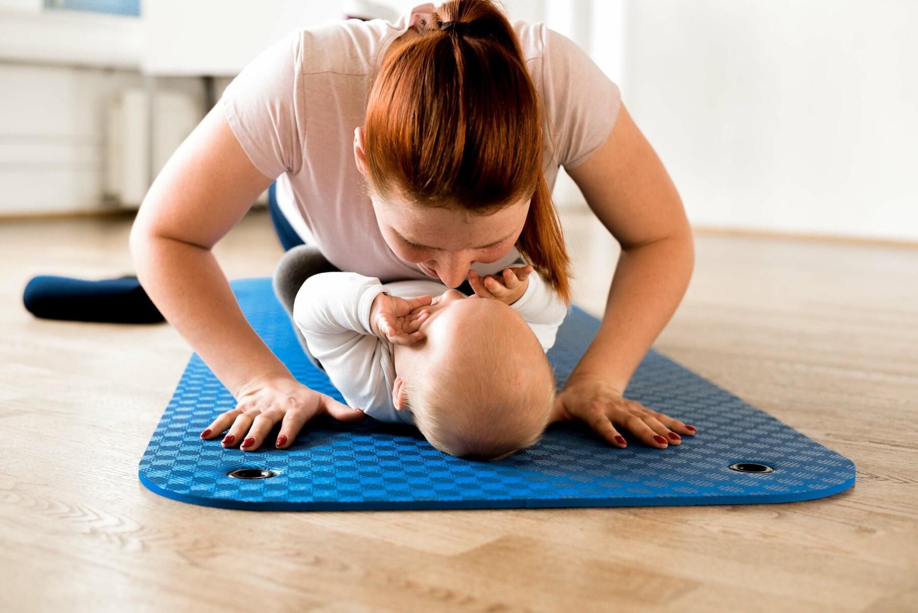 Frau macht Yoga-Übung auf einer blauen Matte und spielt dabei liebevoll mit ihrem Baby im Wohnzimmer.