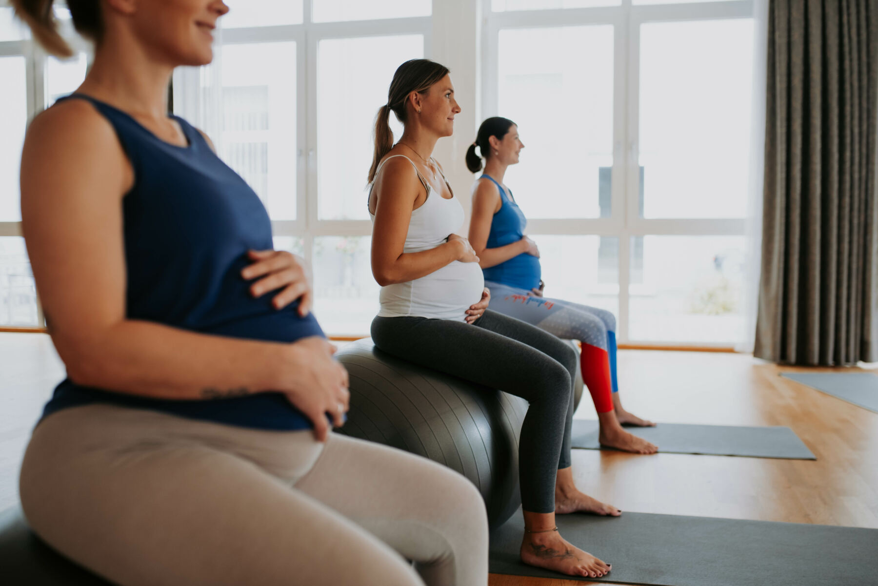 Drei schwangere Frauen sitzen im hellen Studio auf Gymnastikbällen und üben sanfte Bewegung auf Yogamatten.